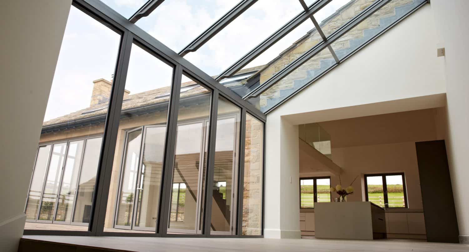 Modern house interior with large floor-to-ceiling glass windows and a glass roof, letting in natural light. The view shows a spacious open plan area with doors leading outside and countryside visible through the windows.