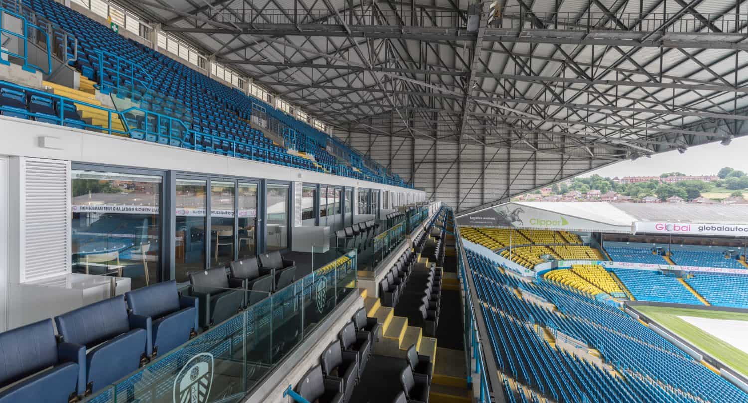 A view inside a large, empty football stadium with blue and yellow seats, bespoke glass solutions in the VIP boxes on the left, and a partially covered metal roof structure overhead. The pitch is visible to the right.
