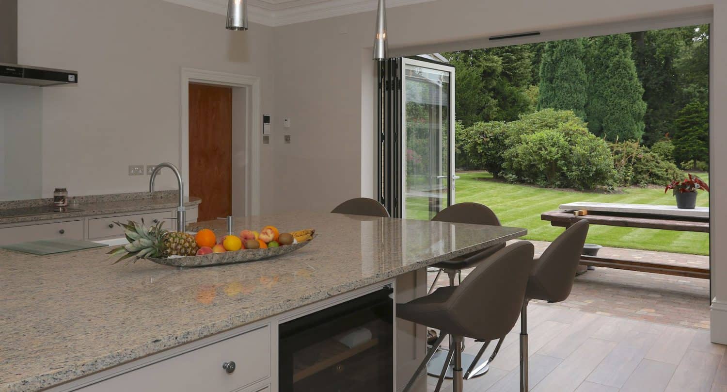 Modern kitchen with a granite island, fruit bowl, and three chairs. Large open glass doors lead to a green backyard with a picnic table and lush trees. Natural light fills the space.