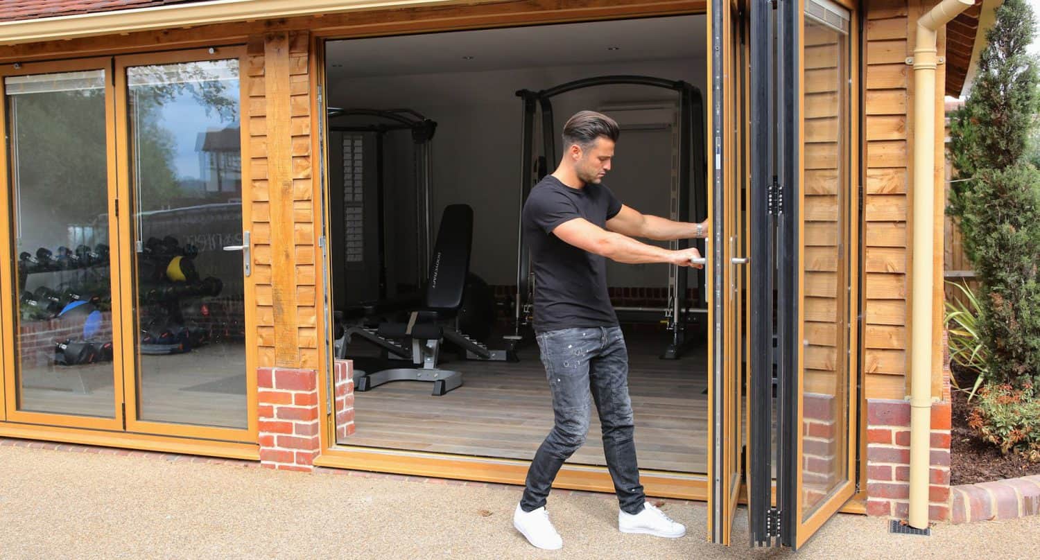 A man in a black t-shirt and jeans opens folding glass doors to a home gym with exercise equipment and weights visible inside. The building has wooden walls and a brick foundation.