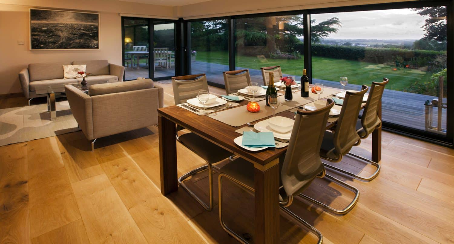 Modern dining room with a set table for six, next to large glass doors overlooking a green garden. The room has wooden floors, contemporary chairs, and a sitting area with two sofas in the background.