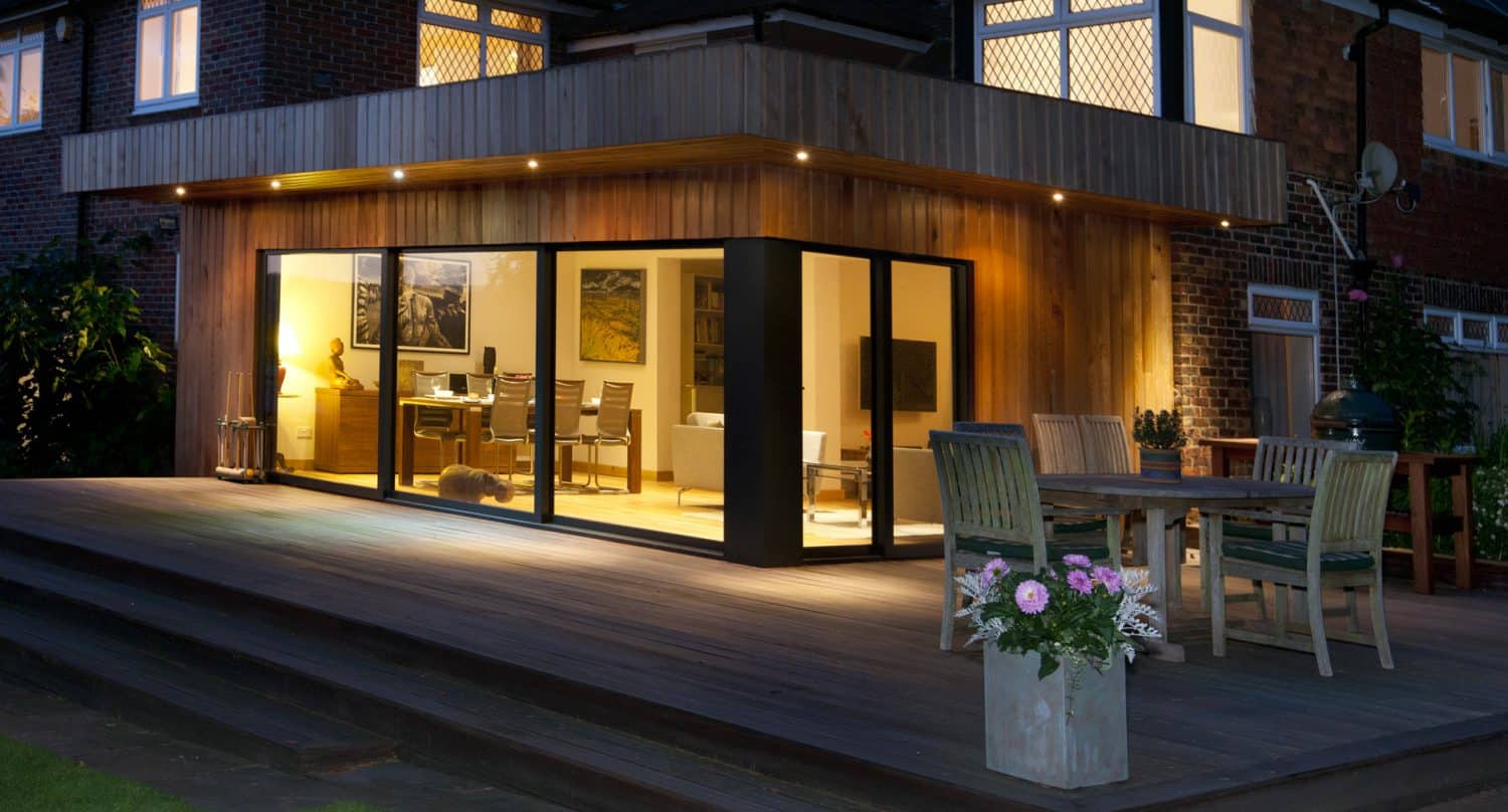 Modern house extension with large floor-to-ceiling glass doors opening onto a wooden deck. Outdoor dining area with wooden table and chairs, potted flowers, and warm indoor lighting visible through the windows.