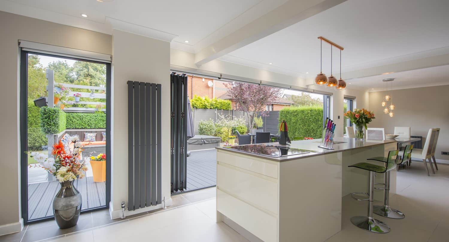 Modern open-plan kitchen and dining area with a large island, bar stools, pendant lights, and sliding doors opening to a landscaped garden and patio. The space is bright and contemporary with neutral tones and fresh flowers.