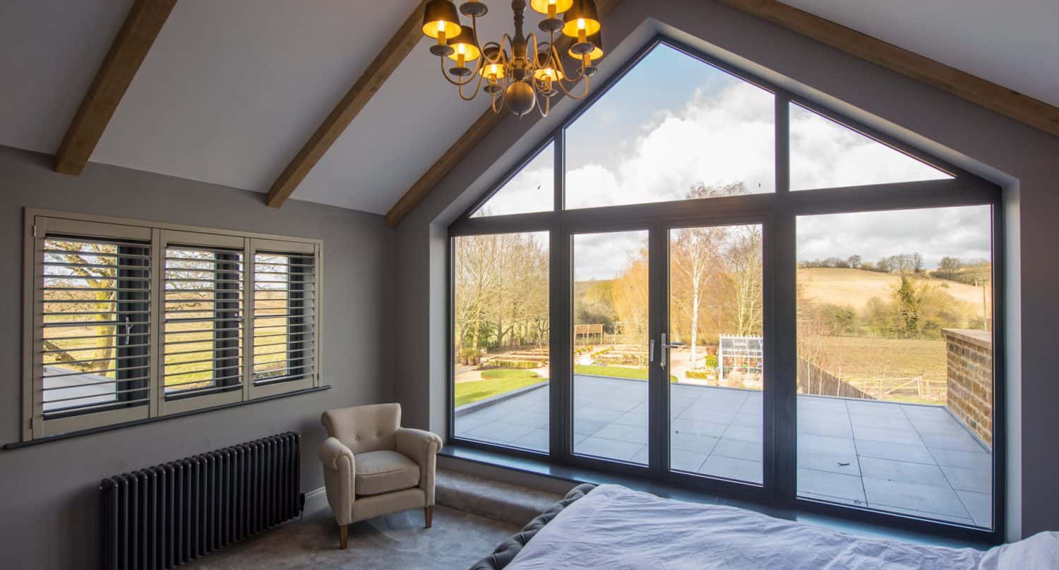 A modern bedroom with vaulted ceilings, exposed wooden beams, a large triangular window and glass doors overlooking a patio and scenic countryside. A beige armchair sits near a window with shutters.