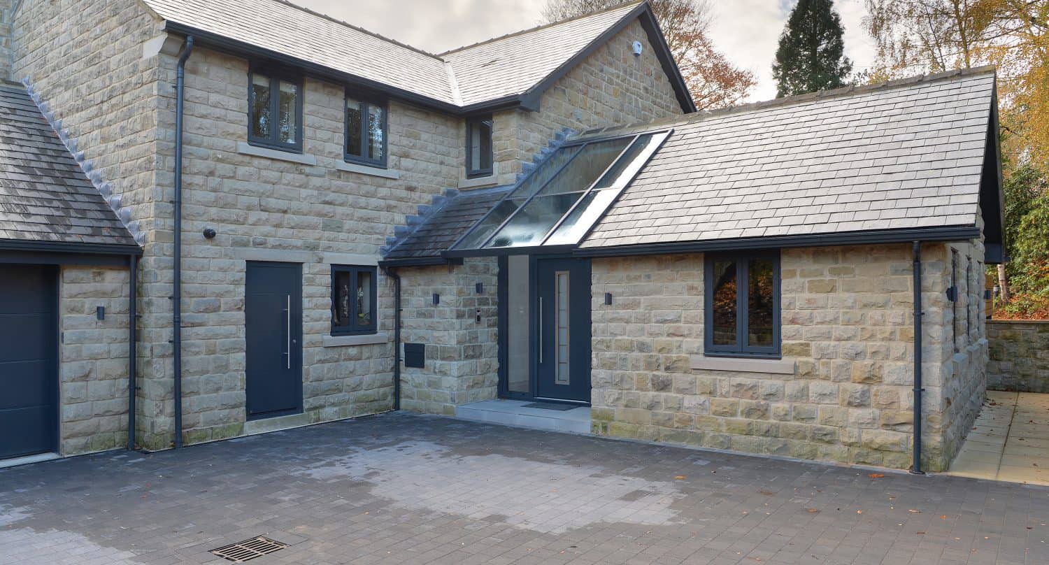 Modern stone house with large windows, a gray front door, and a sloped slate roof. The entrance features bespoke glass solutions with a glass canopy, while the driveway is paved with dark bricks amid trees and autumn foliage.