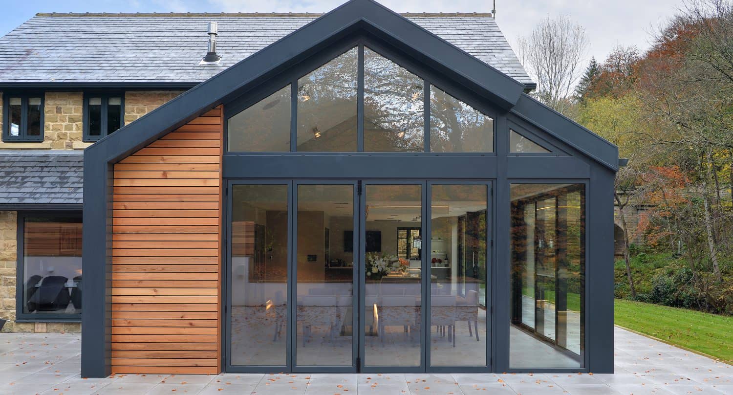 Modern house extension with large sliding doors and windows, black metal framing, and vertical wooden paneling on one side. The bespoke glass solutions open to a spacious patio with trees and greenery in the background.