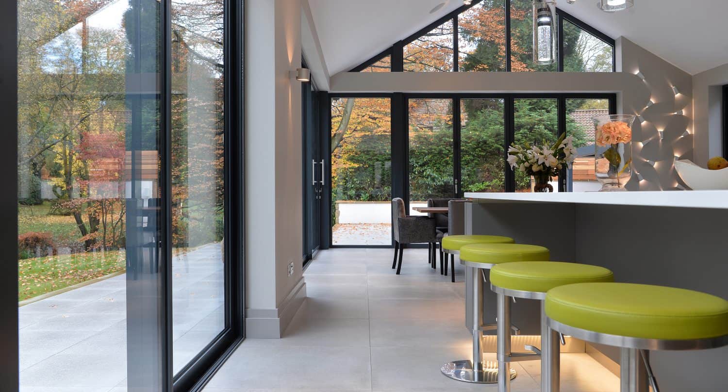 Modern kitchen with large glass windows and bespoke bifold doors overlooking a garden with autumn trees. Green bar stools line a white island, and natural light fills the space. A dining table is visible in the background.