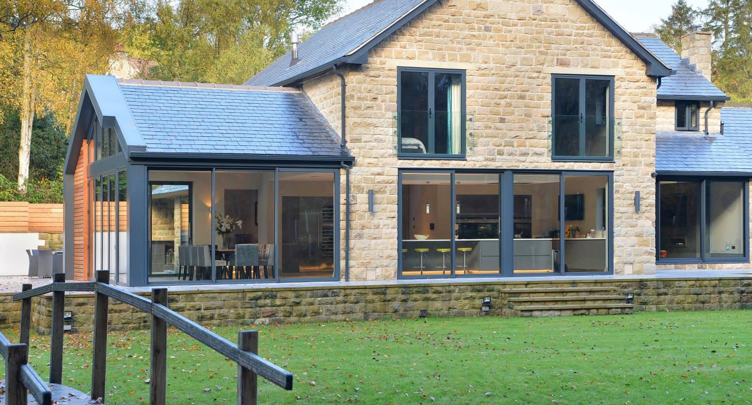 A modern two-story stone house with bespoke glass solutions, including large windows and bifold doors, features a manicured lawn in front and trees in the background. A wooden ramp or walkway leads toward the stunning entry.