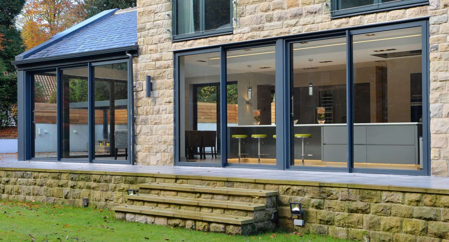 Modern house with large glass sliding doors and stone exterior. Inside, a sleek kitchen with bar stools is visible. Wooden steps lead from the patio to a grassy yard with trees in the background.