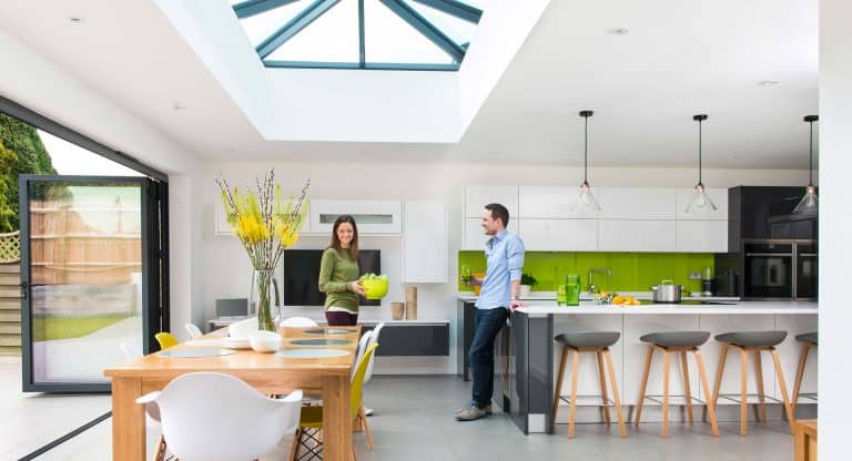 A modern kitchen with a skylight, green accents, and bar stools. A woman holds a green bowl while a man leans on the counter. A wooden dining table with flowers is in the foreground.