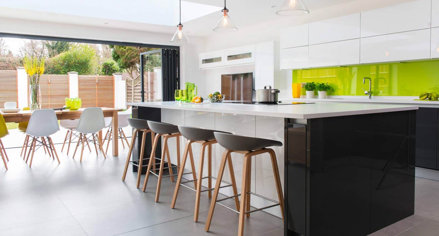 Modern kitchen with a large black and white island, four bar stools, glossy cabinets, and a bright green backsplash. Sliding doors open to a patio with a dining table and chairs, letting in natural light.