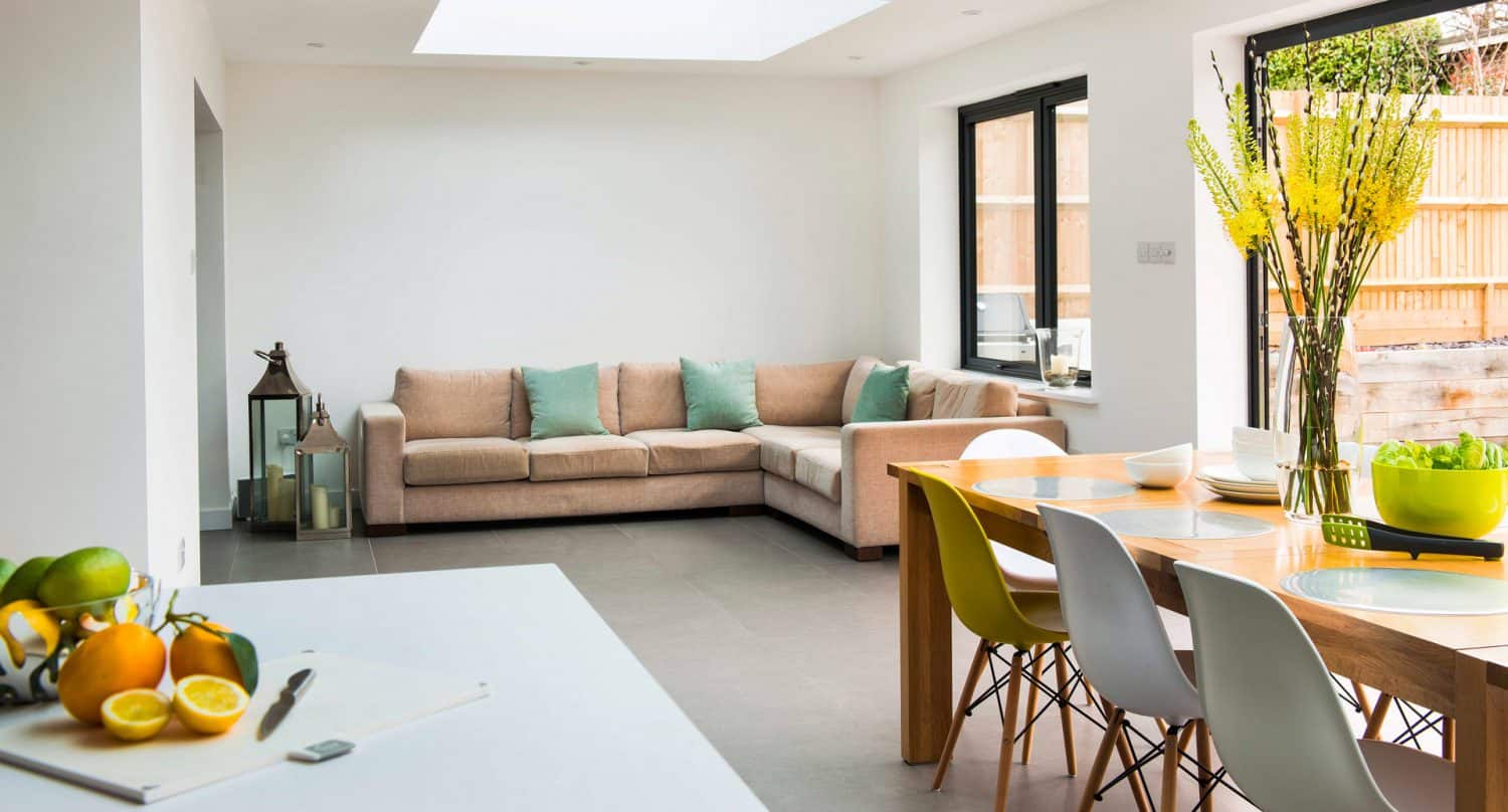 Modern, bright open-plan living and dining area with a beige sofa and pastel cushions, a wooden dining table with multicolored chairs, large windows, and fresh fruit and flowers as decor.