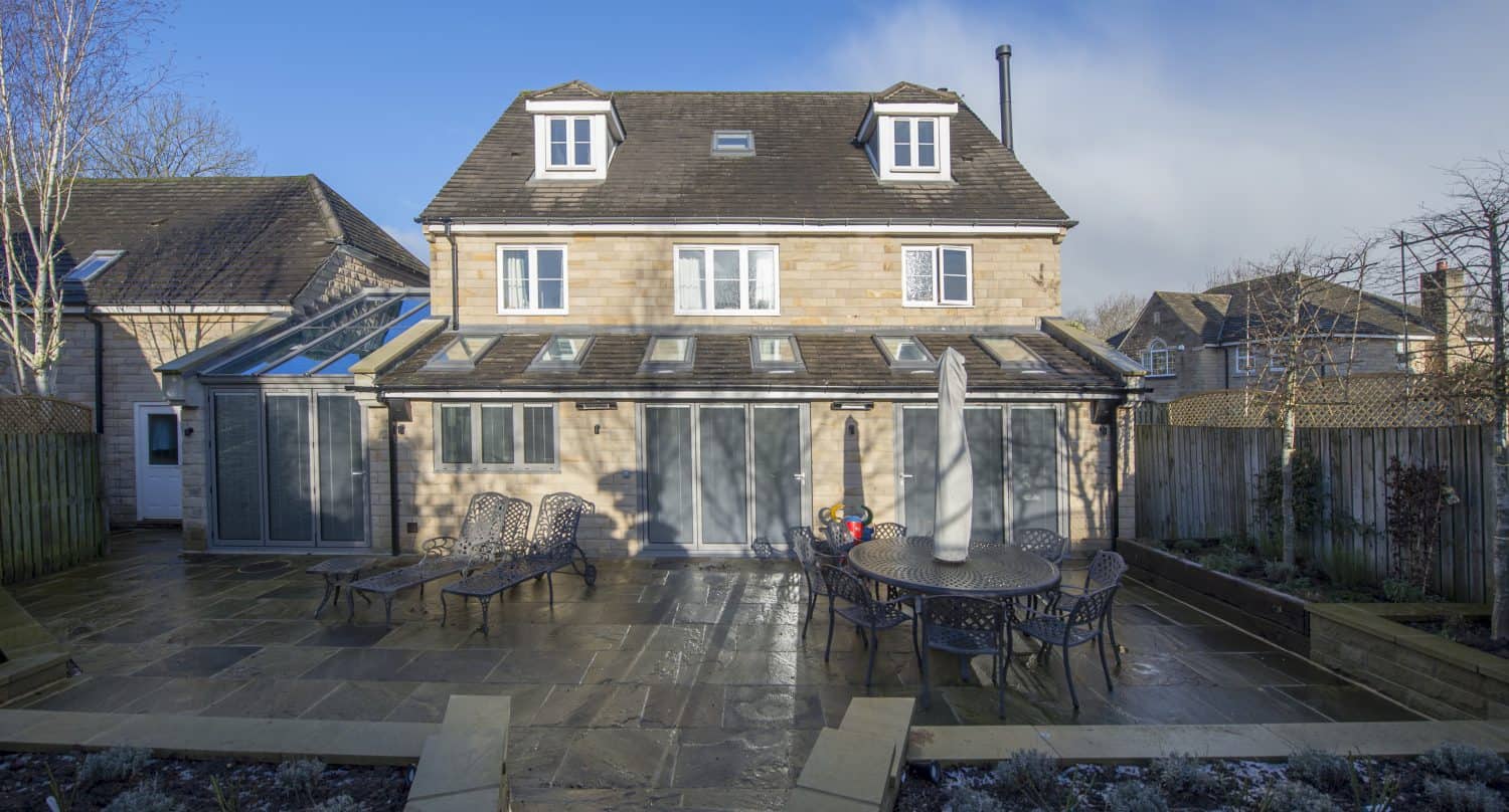 A large, modern stone house with multiple dormer windows features bespoke glass solutions in its sunroom and stylish bifold doors opening onto a spacious patio with lounge chairs and a round dining table. The sky is clear and blue.