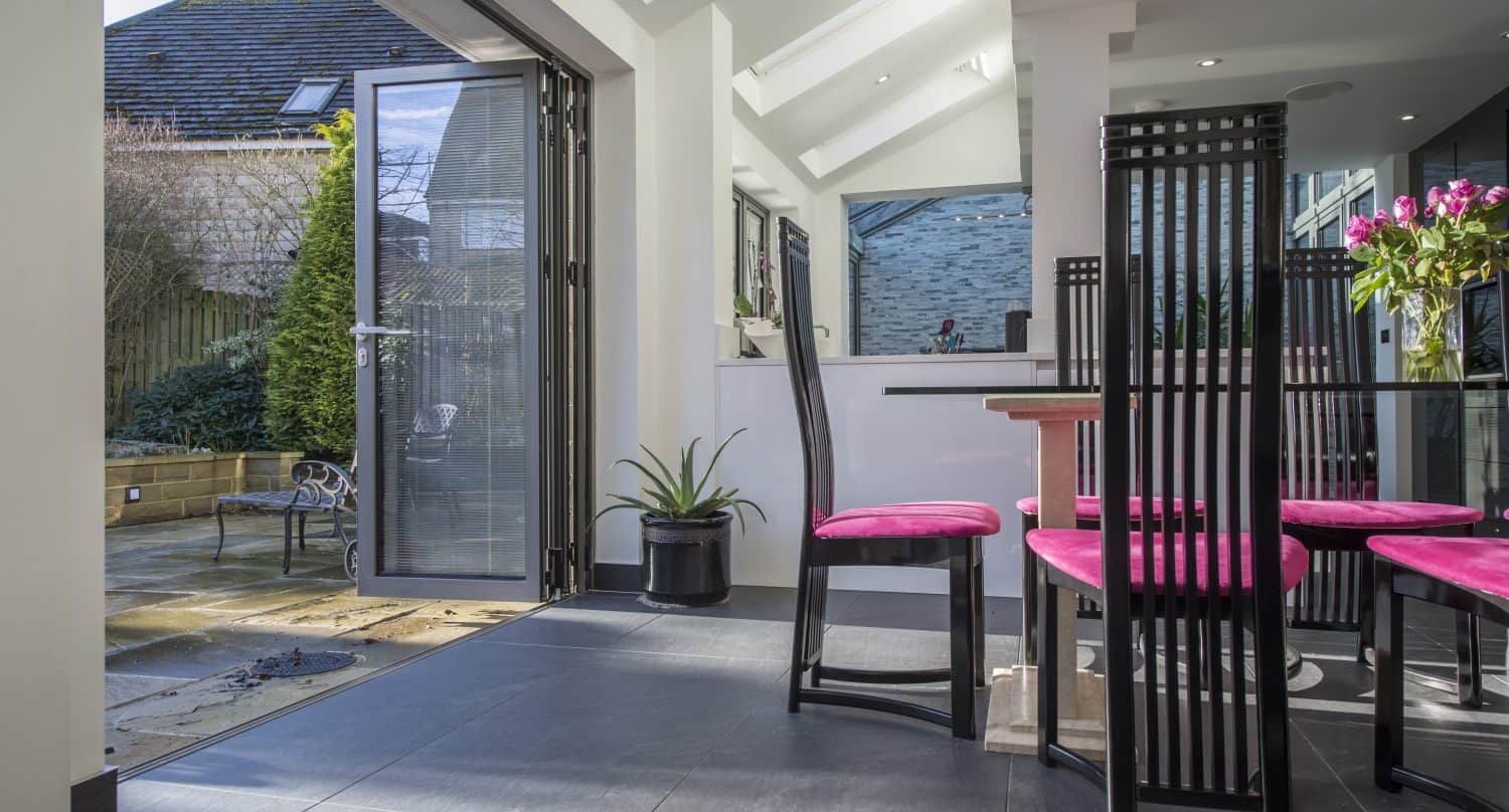 Modern dining area with black chairs featuring bright pink cushions, a glass table with a vase of flowers, and large sliding doors open to a sunny patio with plants and outdoor seating. Natural light fills the space.