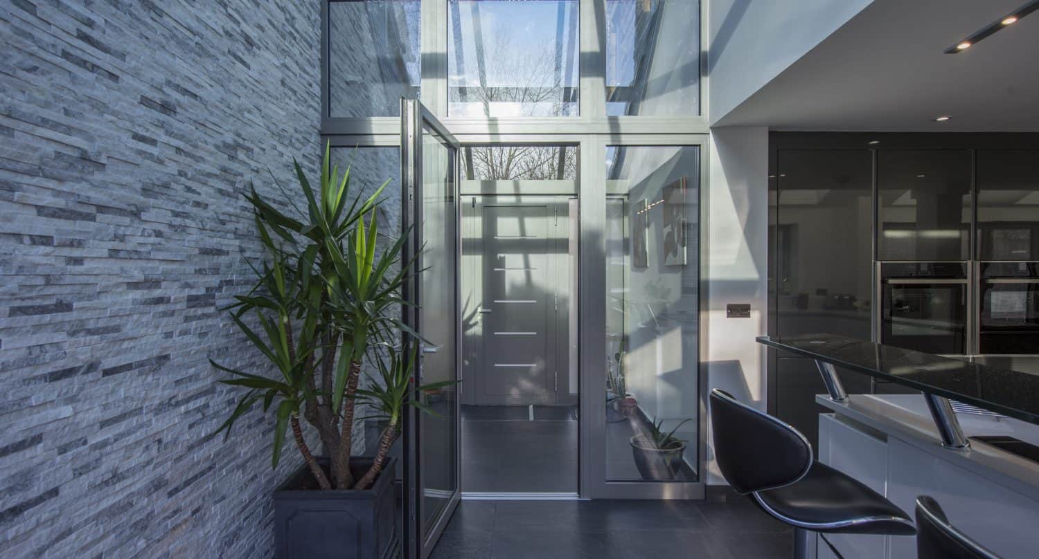 Modern entryway with bespoke glass solutions, including floor-to-ceiling windows and glass front doors, a tall potted plant, gray stone accent wall, and a sleek black-and-white kitchen area with bar stools on the right.