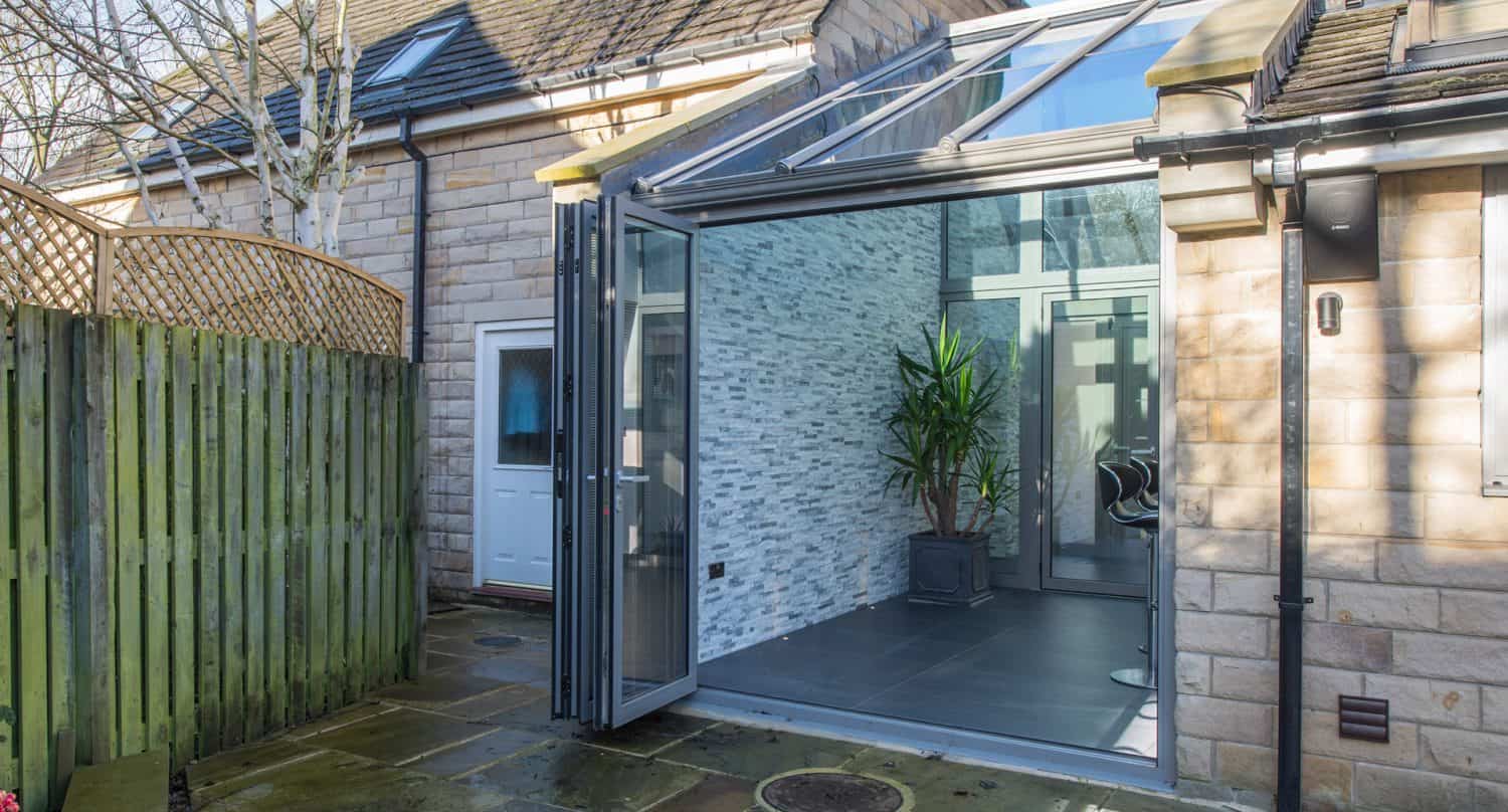 A modern house extension with glass walls and a folding glass door open to a stone patio. Inside, there is a potted plant and gray tiled flooring; outside, there is a wooden fence and stone walls.