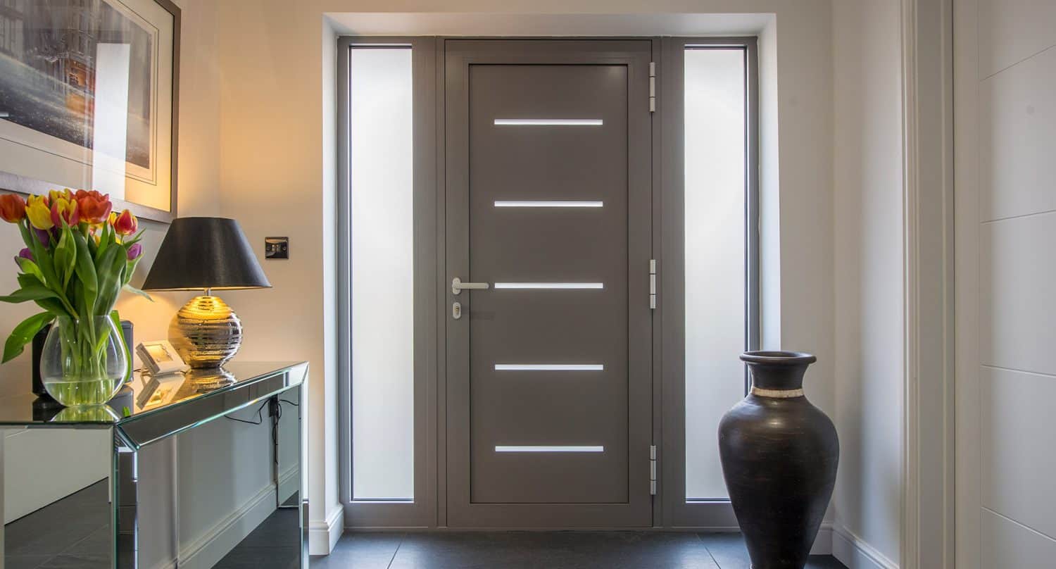 Modern entryway with a gray front door featuring horizontal glass panels, flanked by narrow windows. A large ceramic vase sits on the right, while a table with flowers, a lamp, and framed photos is on the left.