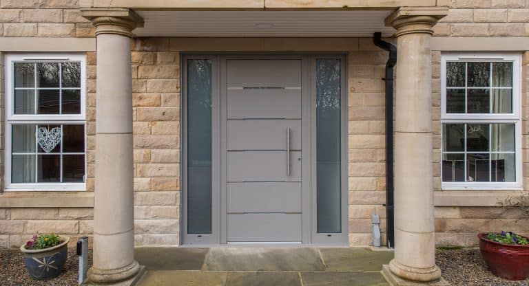 Modern grey front door with horizontal lines, flanked by narrow frosted glass panels, set in a stone exterior with two large columns. Windows with white frames and potted plants are on either side of the entrance.