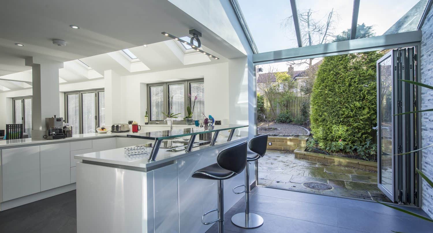 Modern kitchen with white cabinets and a breakfast bar with two black stools; large bifold doors open to a patio and garden, flooding the space with natural light. Skylights and windows complete the bright, airy atmosphere.