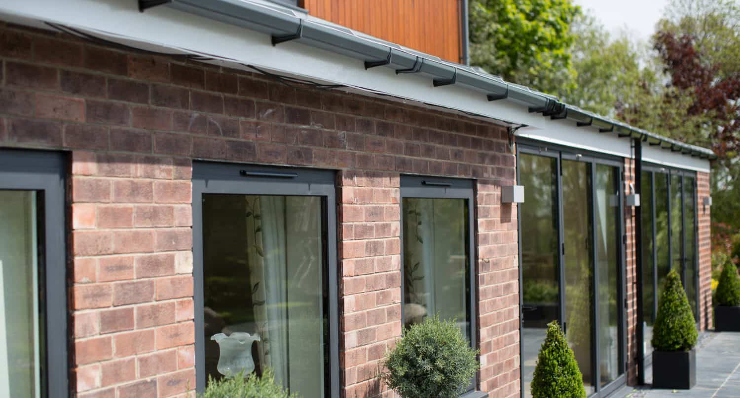 Modern brick house with large windows, glass roofing, and sliding doors, potted plants along a stone pathway, and greenery visible in the background.