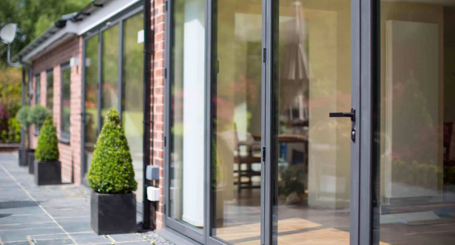 Modern house exterior with large glass sliding doors, brick walls, and neatly trimmed potted plants along a tiled patio. The doors are open, revealing a glimpse of the interior and an umbrella.