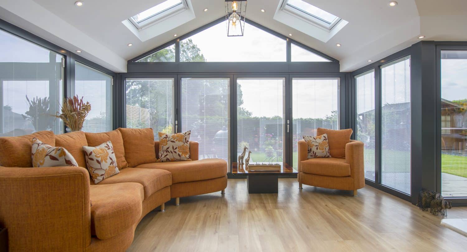 A bright sunroom with large windows, a light wood floor, two orange sofas with patterned cushions, skylights, and a glass table holding decorative items, features bifold doors opening to a green garden.