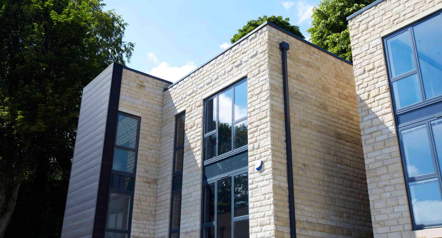 Modern building with light stone brick walls, large windows, and black metal trim, surrounded by green trees under a clear blue sky with a few clouds.
