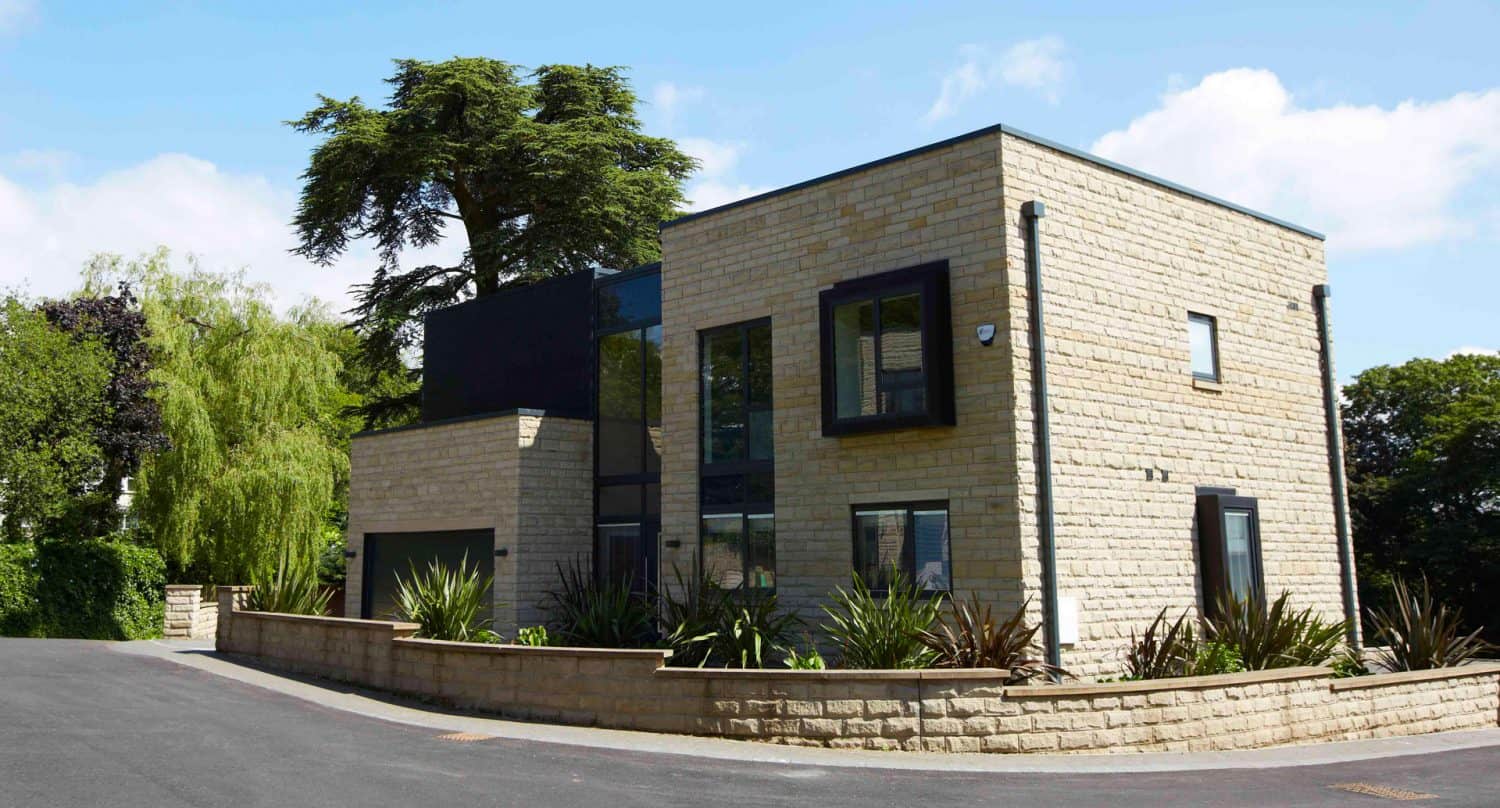 Modern two-story house with light stone exterior, large windows, black trim, and a landscaped front garden, situated on a corner lot with trees and blue sky in the background.