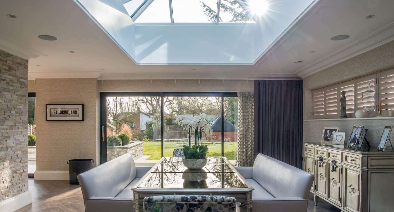 A modern dining area with a glass ceiling, mirrored table, and cushioned benches is filled with natural light from a large skylight and sliding glass doors that open to a green garden with trees and a gazebo.
