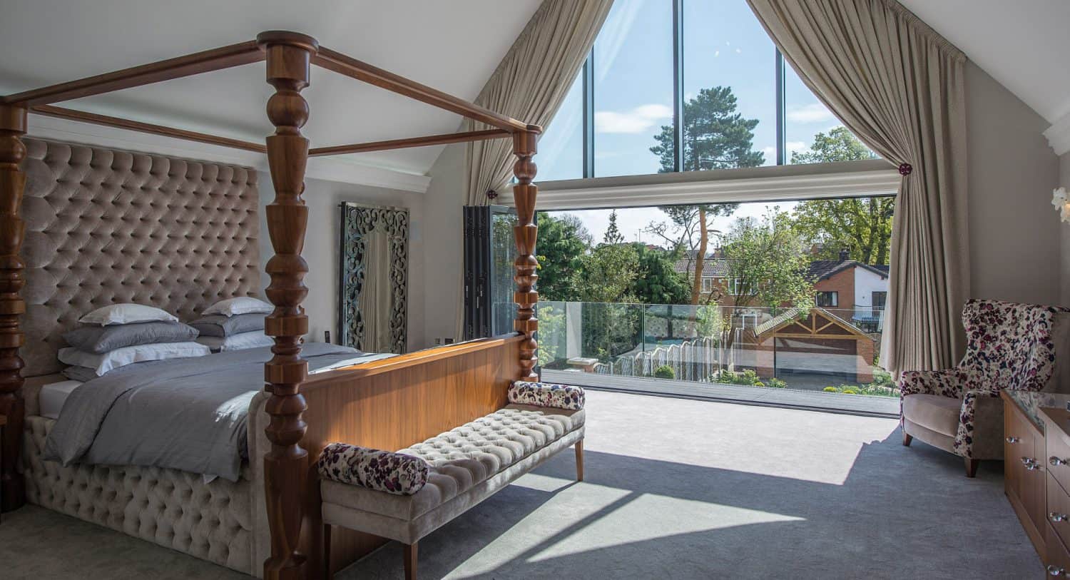 Spacious bedroom with a four-poster bed, upholstered bench, and armchairs. Large floor-to-ceiling windows with drapes open to a balcony, letting in natural light and offering views of trees and houses outside.