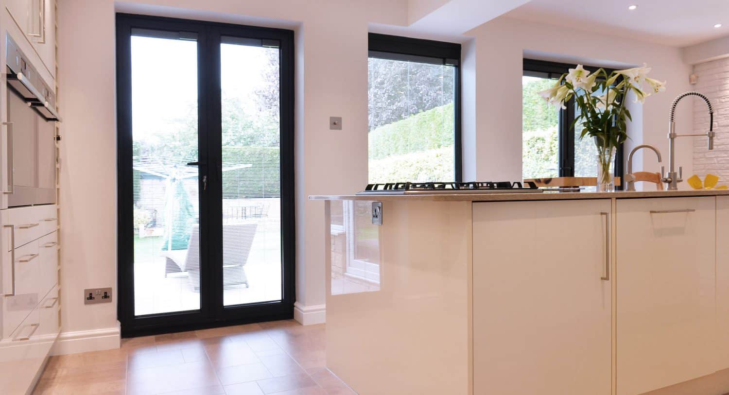 Modern kitchen with glossy white cabinets, an island with a built-in stove, tall black-framed glass doors, large windows, and a vase of white lilies on the countertop. Outdoor greenery is visible through the doors.