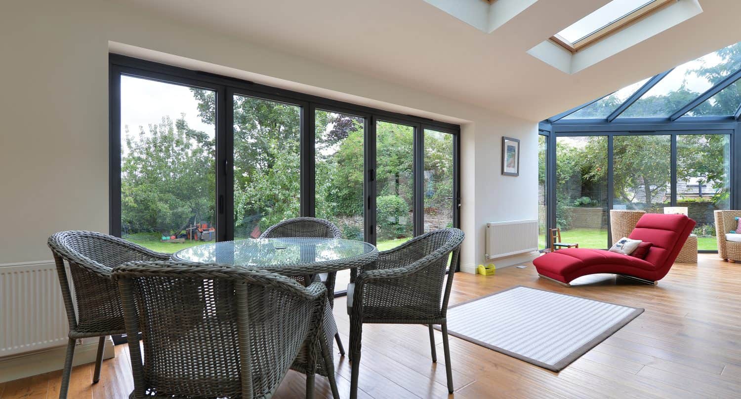 A bright sunroom with bespoke glass solutions, including skylights and bifold doors, features a round glass dining table with four wicker chairs, a red lounge chair, and views of a green garden outside.