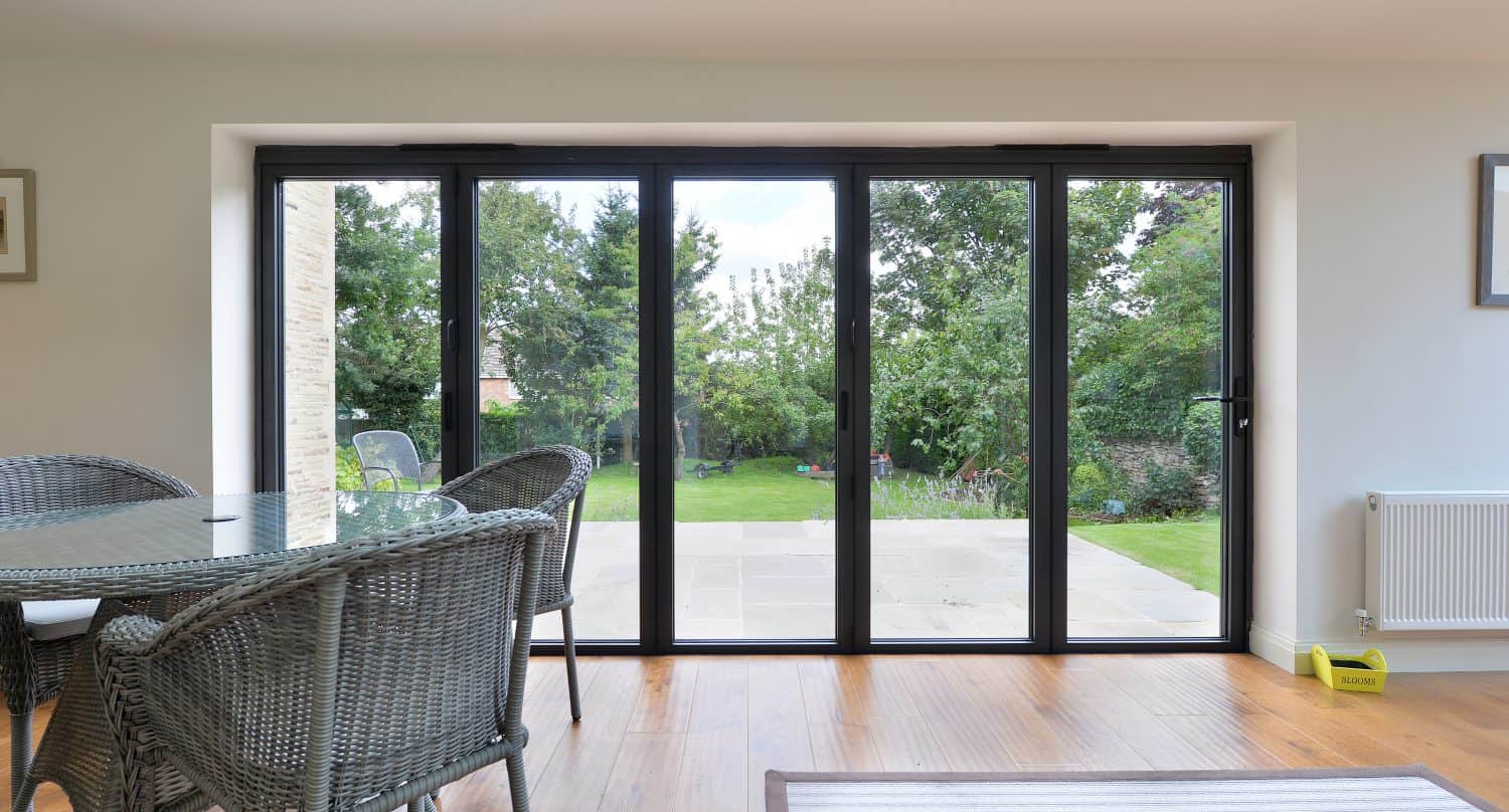 A modern dining area with wicker chairs and a glass table beside large sliding glass doors, crafted with bespoke glass solutions, offering a view of a green garden and patio outside. Natural light fills the room, highlighting the wooden floor.