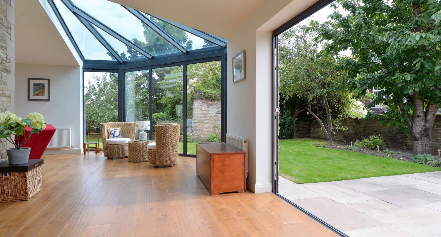 A bright sunroom with bespoke glass solutions features large glass windows and a glass roofing, wooden floor, wicker chairs, and open doors leading to a green garden with trees and a stone wall.