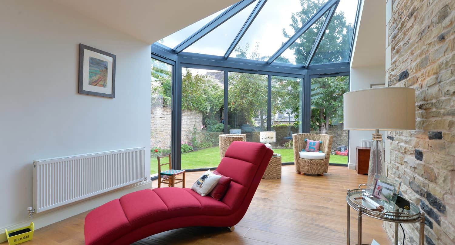 A modern sunroom with bespoke glass solutions, large windows and ceiling, featuring a red chaise lounge, wicker chairs, wooden floor, stone accent wall, and views of a green garden outside.