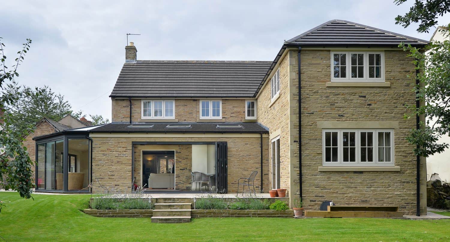 A modern two-story house with tan brick exterior, large windows, a glass sunroom extension on the left, and a well-manicured green lawn with steps leading to a patio area.