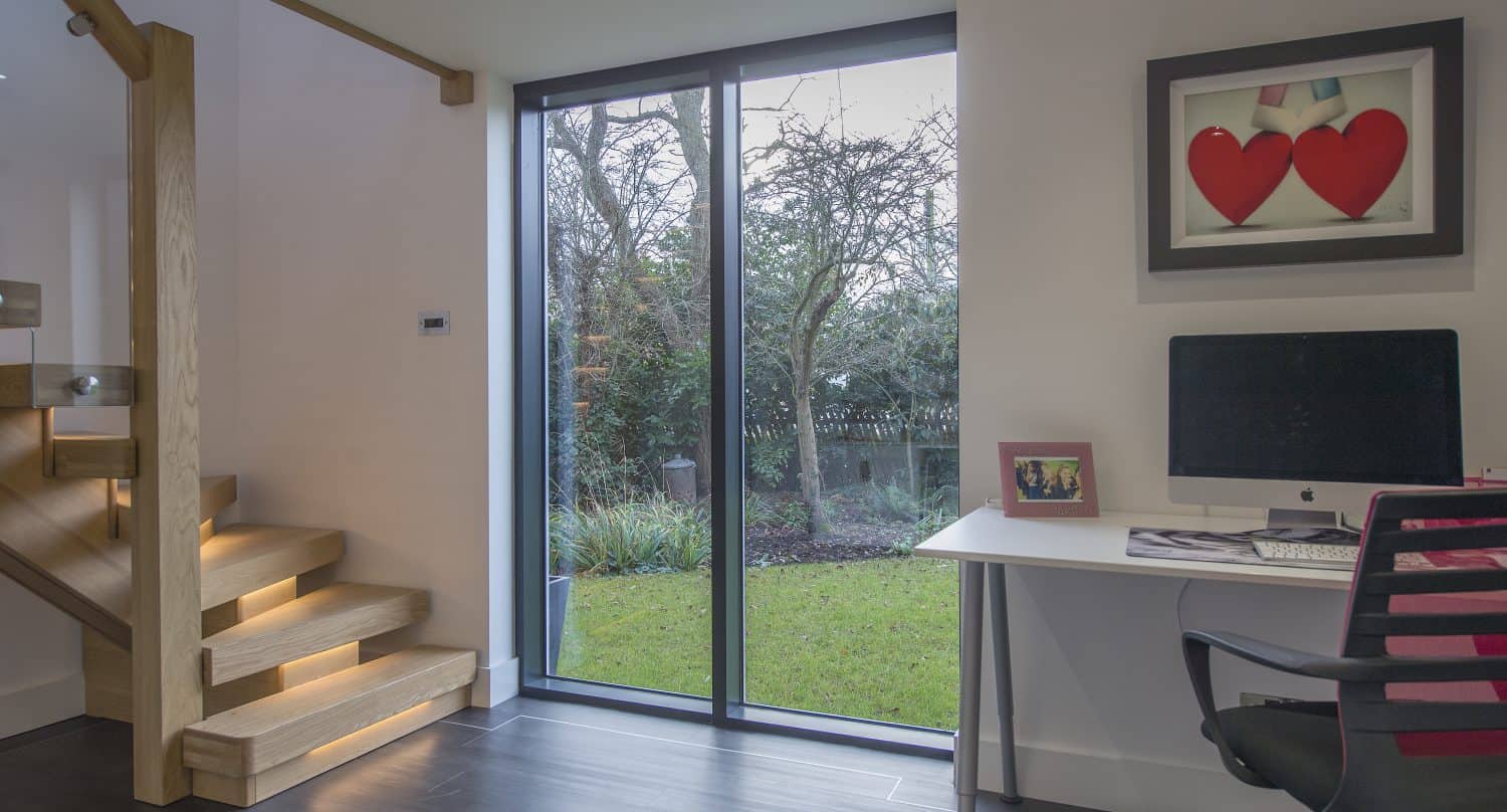 Modern home office with large sliding doors overlooking a garden, wooden staircase with built-in lights, desk with computer, chair, photo frame, and wall art featuring two red hearts.