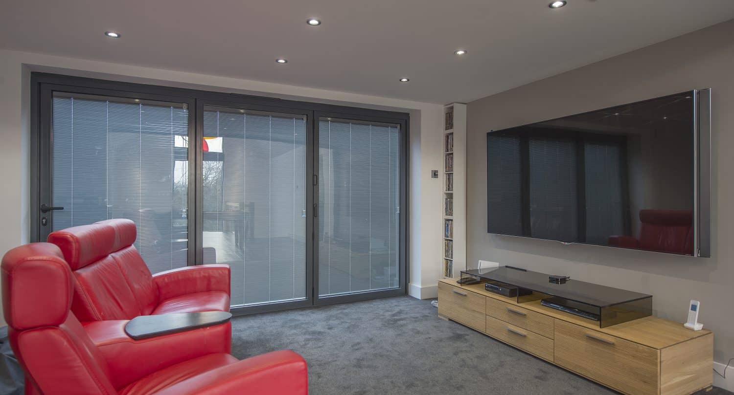 A modern home theater room with red leather seats, a large wall-mounted flat-screen TV, light wood media console, and floor-to-ceiling sliding doors with blinds. Recessed ceiling lighting enhances the spaces sleek look.