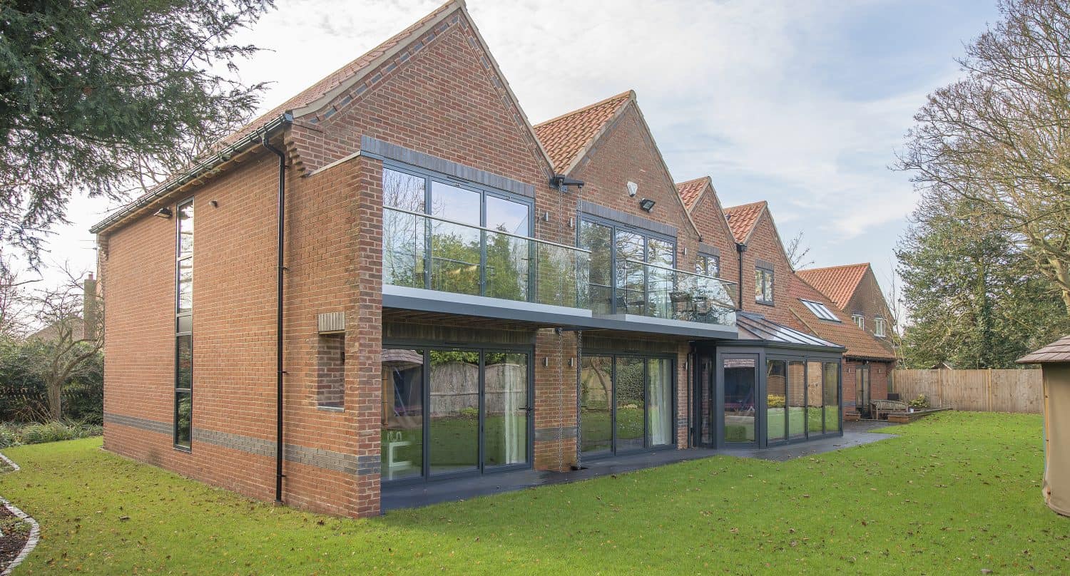 A modern two-story brick house with large glass windows, bifold doors opening to a balcony, surrounded by a green lawn and trees under a partly cloudy sky.