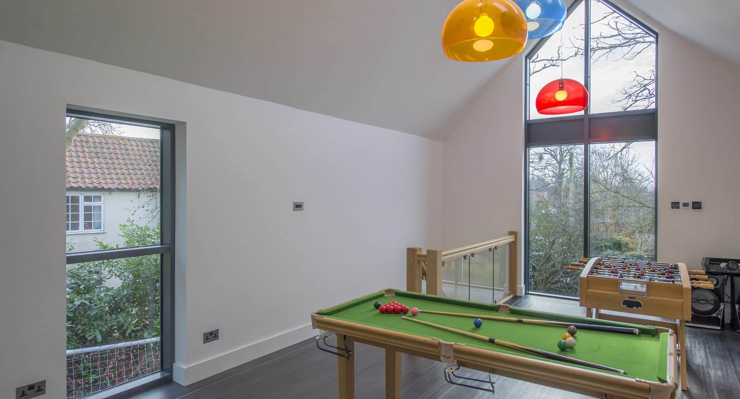 A modern game room with a mini pool table and foosball table, large windows and sliding doors letting in natural light, and colorful pendant lights hanging from a high, sloped ceiling. Trees and a neighboring house are visible outside.