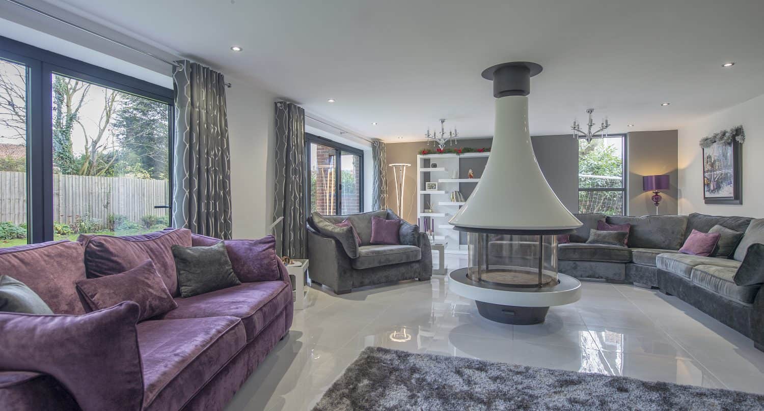 Modern living room with large windows, gray and purple sofas, a central white suspended fireplace, plush rug, and contemporary decor. Bespoke glass solutions like sliding doors let natural light fill the spacious, open room.