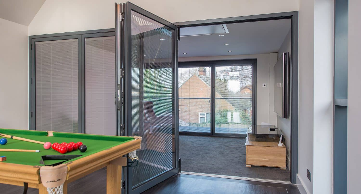A modern room with large folding glass doors, leading to a carpeted area with a TV stand. In the foreground is a pool table with cues and balls. Large windows provide views of neighboring houses and trees.