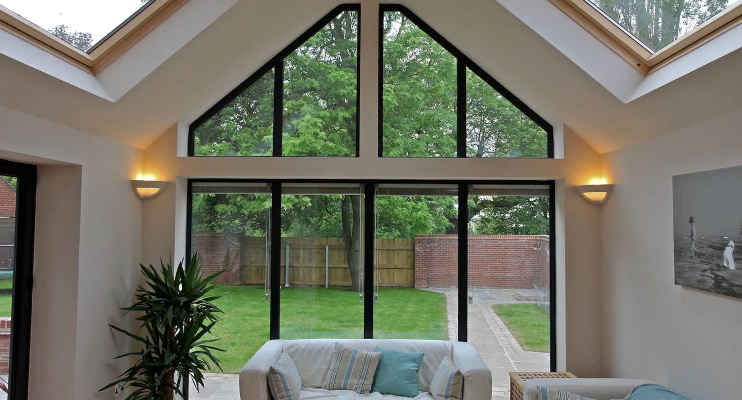 Bright living room with large triangular and rectangular windows facing a green garden. White sofas, potted plant, wall lights, and a coastal-themed picture decorate the space. Skylights let in additional natural light.