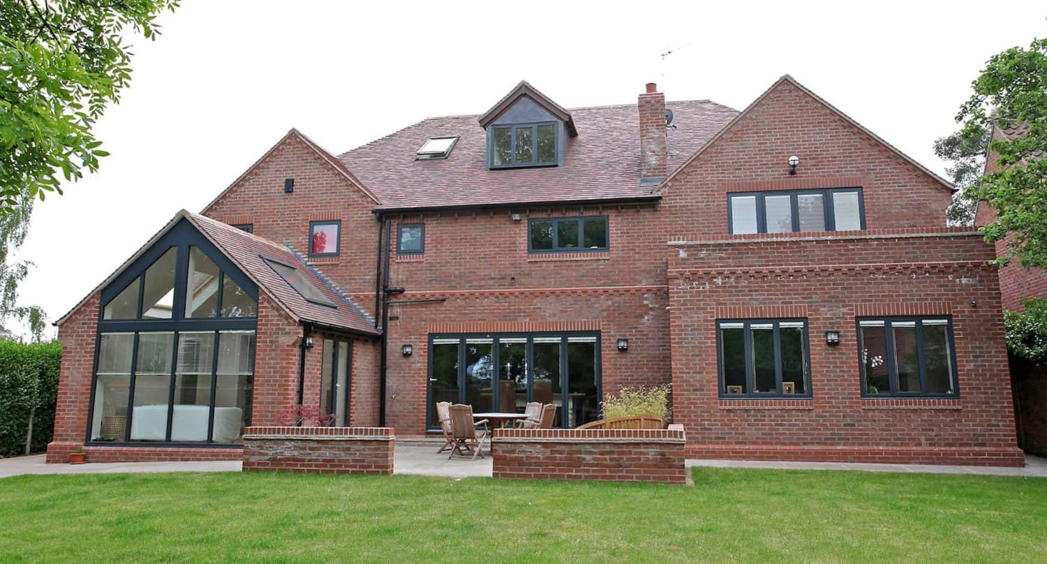A large, modern red-brick house with multiple windows, a glass-enclosed sunroom, and a patio with outdoor furniture, set in a grassy backyard.