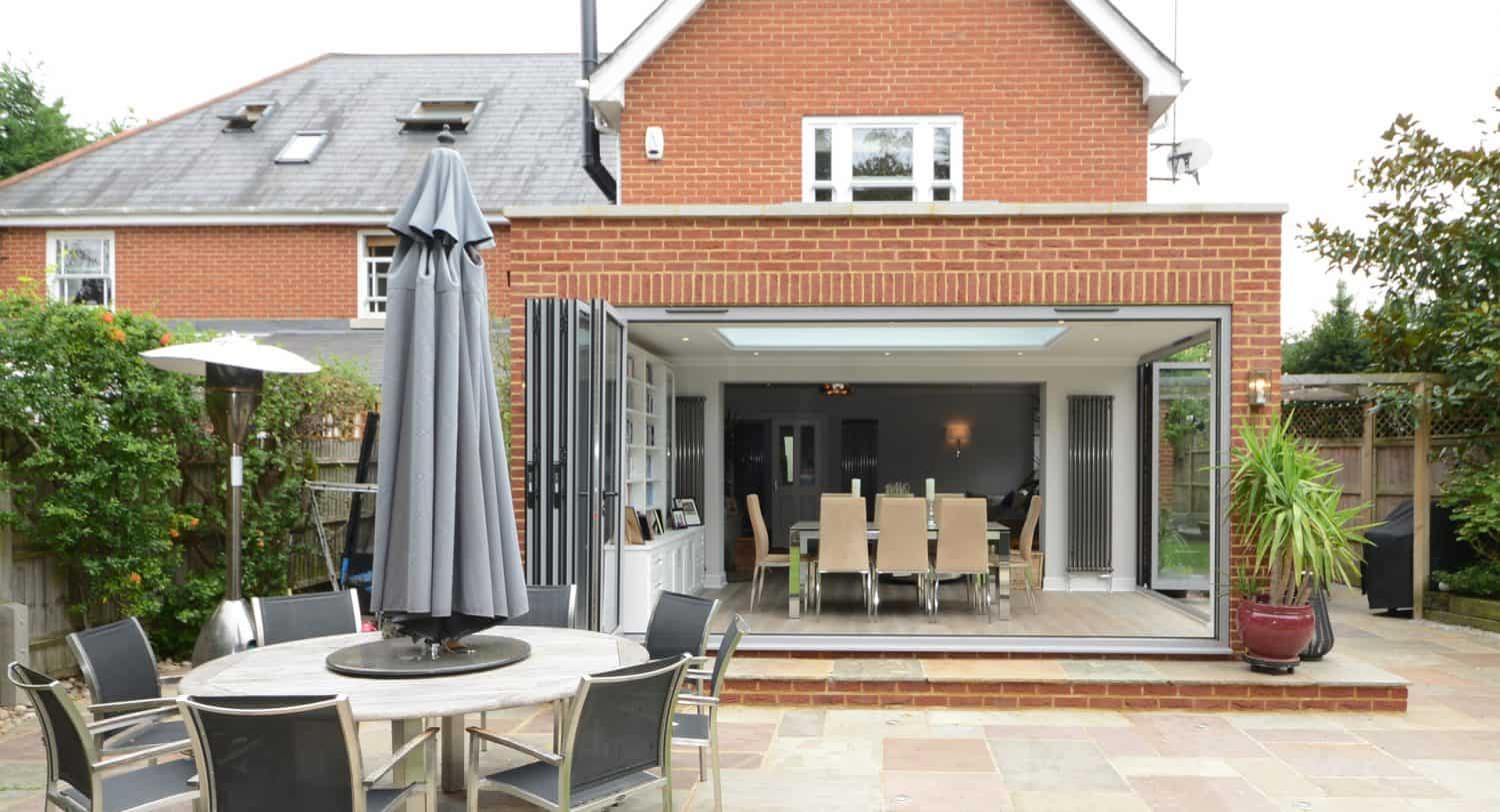 A modern brick house with large, open glass doors leading to a dining area. Outside, there’s a patio with a round table, black chairs, and a closed umbrella, surrounded by greenery.