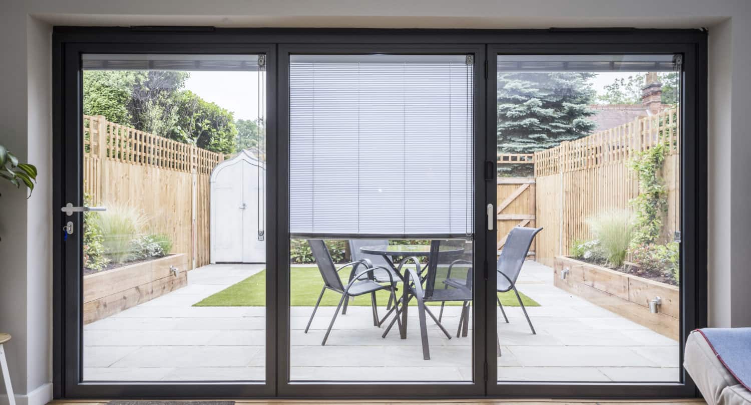 A view through large bifold doors reveals a modern patio with a table, four chairs, potted plants, a grassy area, wooden fences, and a small shed at the back—showcasing bespoke glass solutions for seamless indoor-outdoor living.