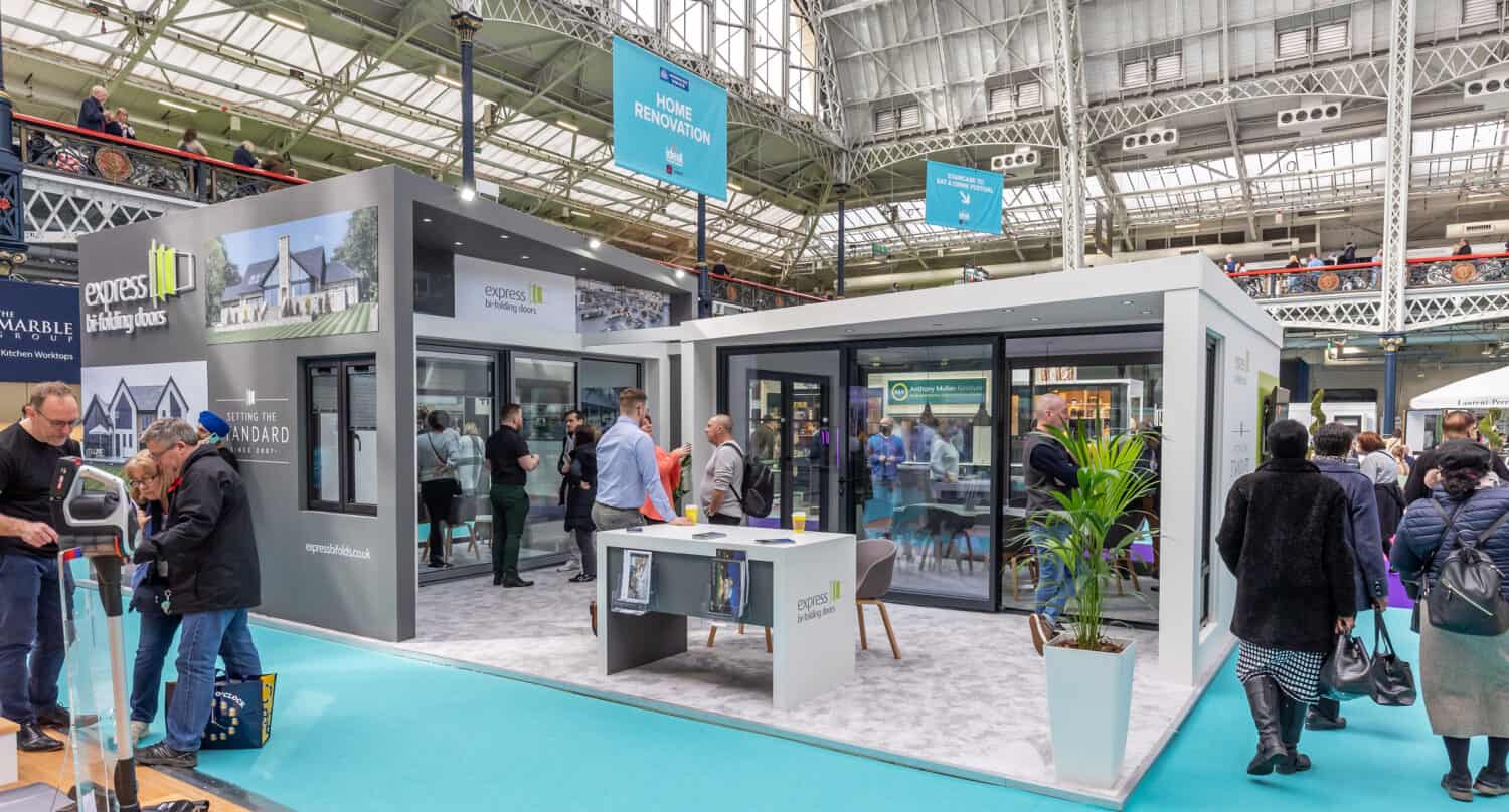 A trade show booth displays modern glass doors, bifold doors, and windows, with people walking around, talking, and exploring the exhibit inside a large, well-lit convention hall.