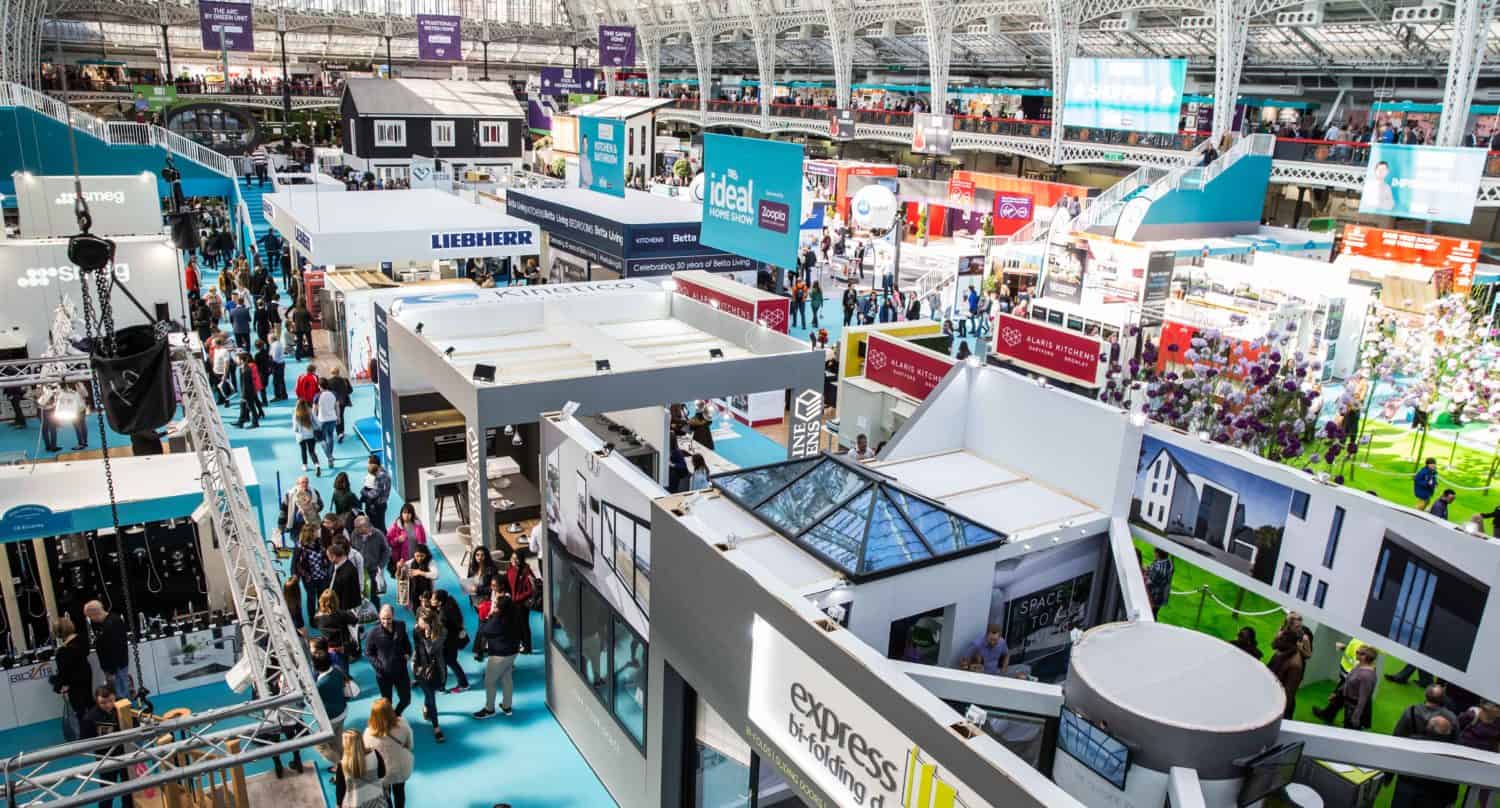 Large indoor exhibition hall bustling with crowds exploring home and garden brands. The space is brightly lit, boasting a high arched glass ceiling that showcases bespoke glass solutions among the stunning booths and displays.