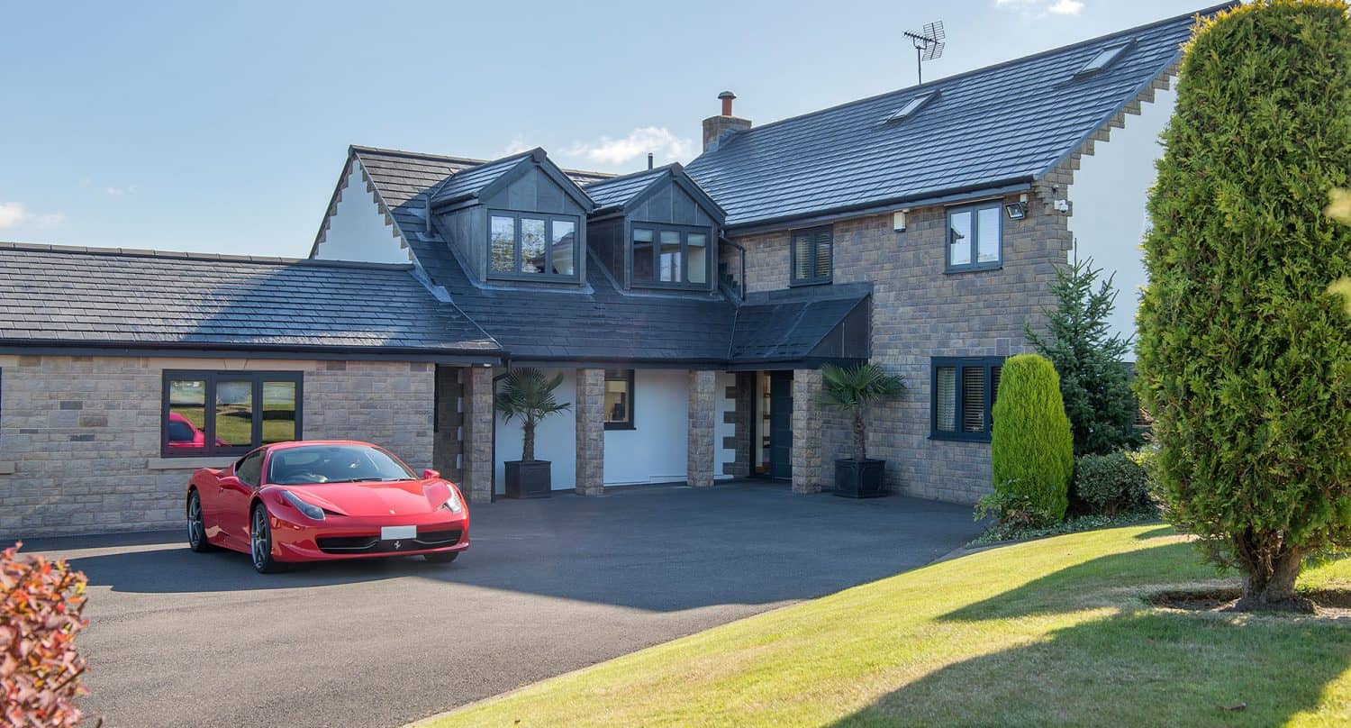 Modern two-story stone house with a dark roof and large windows, featuring sleek bifold doors, a red sports car parked in the driveway, and a well-maintained front lawn with shrubs and trees.