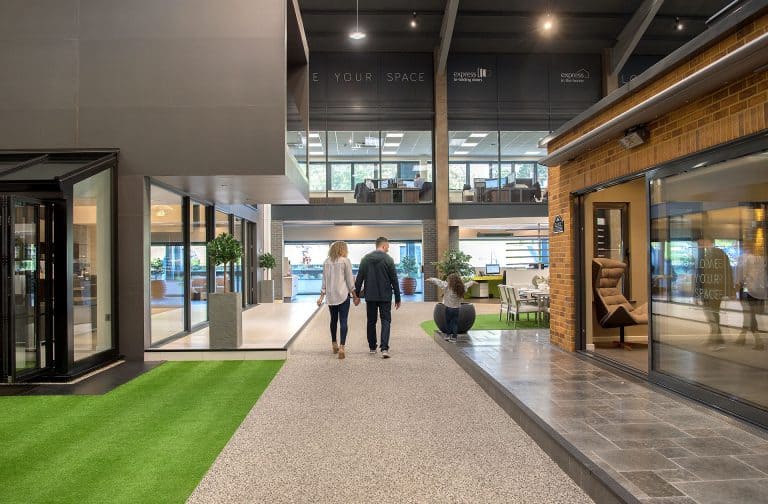 A man and woman walk hand-in-hand through a modern, open indoor space with artificial grass, large windows, and office areas. Sliding doors lead to the area, while another person sits on a bench nearby. The atmosphere is bright and contemporary.